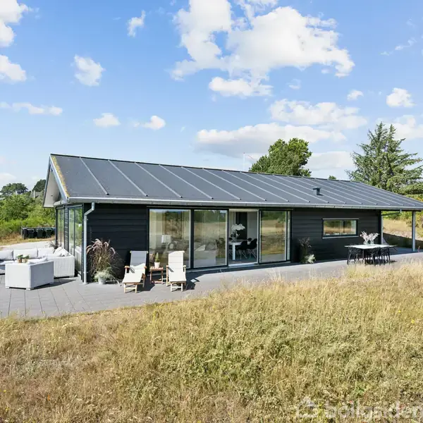 Et moderne sort træhus med store vinduer står på en terrasse. Husets terrasse er omgivet af tørret græs og grønne træer i baggrunden under en blå himmel med skyer.