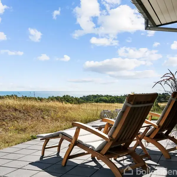 To træstole står på en terrasse med fliser. De vender ud mod en udsigt over en mark og havet i baggrunden. Solrig dag med blå skyhimmel.