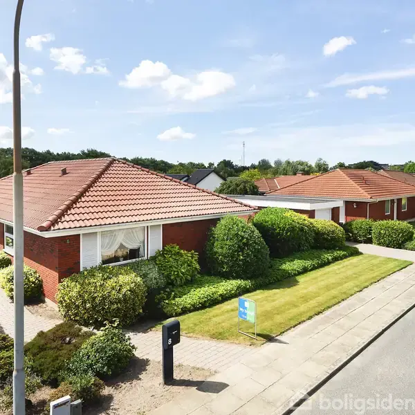 En murstensvilla med rødt tegltag står i et villakvarter. Villaen er omkranset af en velplejet have med buske. En asfalteret vej løber forbi huset.