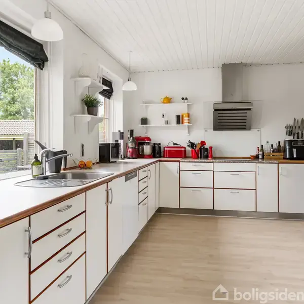 Et lyst køkken med hvide skabe og trækanter. Køkkengrej og apparater, herunder en brødrister og kaffemaskine, står på bordpladerne. Store vinduer giver udsigt til haven.