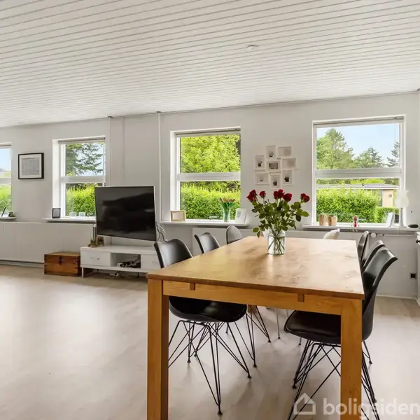 Et lyst rum med et stort spisebord og seks sorte stole i centrum. På bordet står en vase med røde blomster. Rummet har fem vinduer med udsigt til grønne træer. En fladskærms-tv står til venstre.