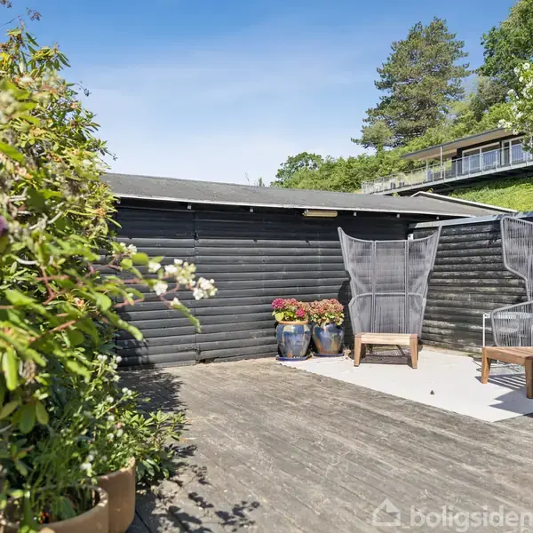 En terrasse med træstole og læskærme står foran et sort malet skur. Omkring er der blomster i potter og træer i baggrunden under en klar blå himmel.