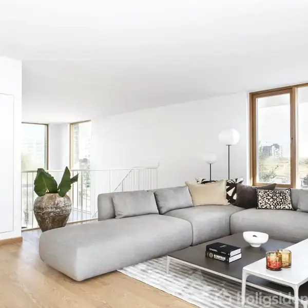 Grå sofa står i en lys stue med store vinduer. En potteplante og en gulvlampe dekorerer rummet. Rummet har parketgulv og hvide vægge, der skaber en minimalistisk stil.