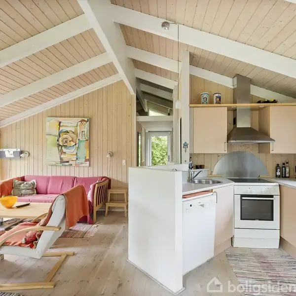 Stue i et sommerhus med træloft og -vægge. En rød sofa med farverige puder står ved et vindue; ved siden af er der en lænestol. Et åbent køkken har komfur og køkkenskabe.