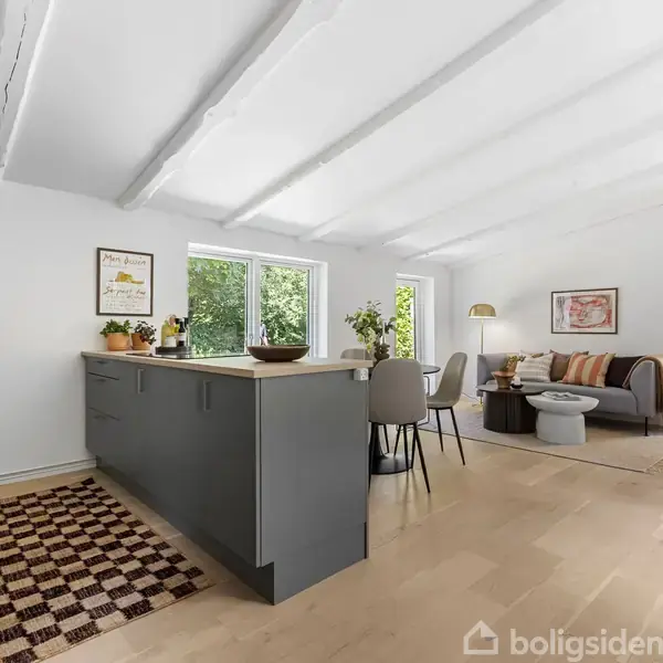 Køkkenalrum med gråt køkkenø med skåle og planter. Spisebord med stole og sofa i lyst, rummeligt rum. Store vinduer giver udsigt til grønne træer. Ruder skaber lys, rolig atmosfære.