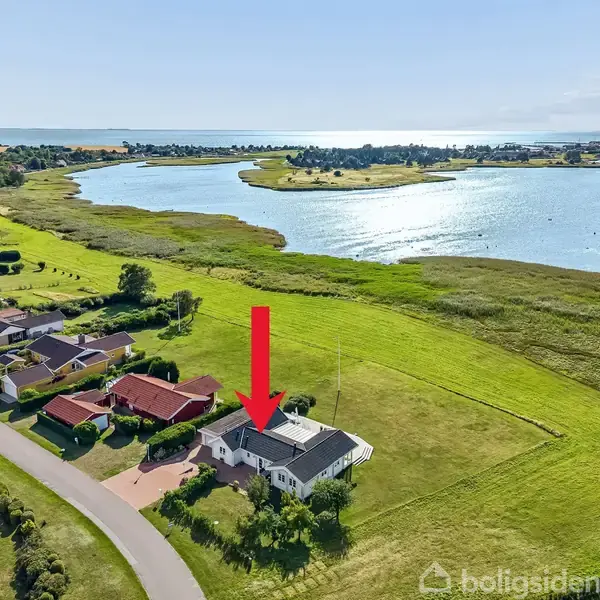 Et hus markeret med en rød pil ligger i et grønt område nær en bugt. Omgivet af græsplæner og flere huse på en vej.