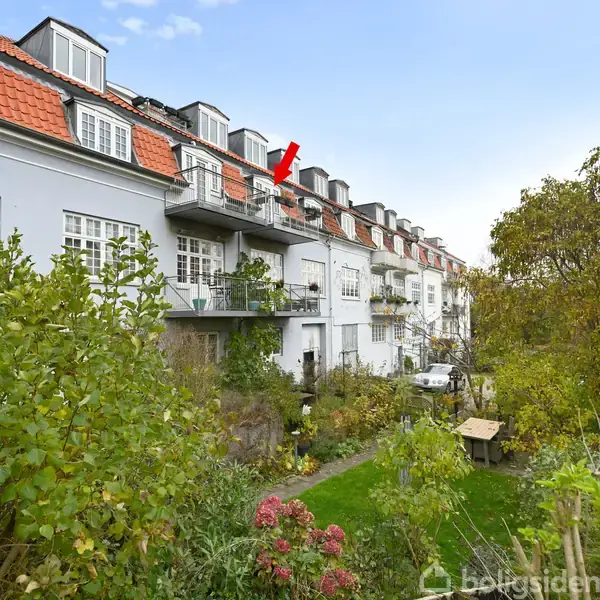 En række hvide byhuse med røde tage har altaner ud mod en frodig have. En rød pil peger mod en specifik altan. Græsplæne og træer fylder haven.