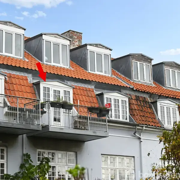 Bygning med grå facade og rødt tegltag har flere altaner. En rød pil peger på en specifik balkon. Grønne træer i forgrunden og blå himmel i baggrunden.