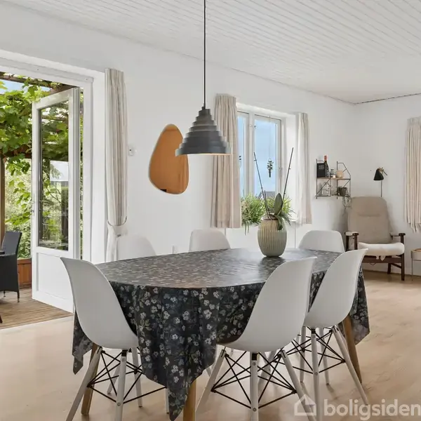 Spisebord med blomsterdug og seks hvide stole. Ovenfor hænger en lampe. I baggrunden åbne døre til terrasse med planter, og store vinduer med udsigt til haven.