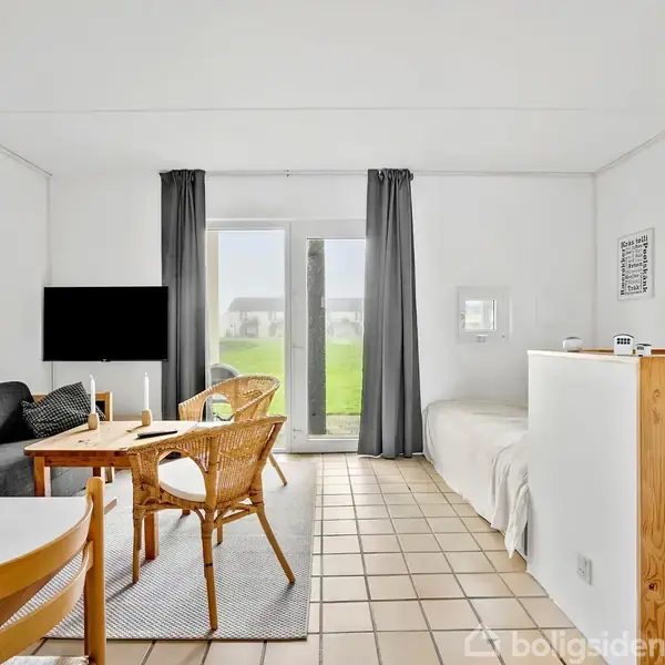 Stue med sofa, tv og trækbord. Står på flisegulv, ved siden af seng. Store vinduer med gardiner. Lys og minimalistisk indretning med grøn udsigt udenfor.