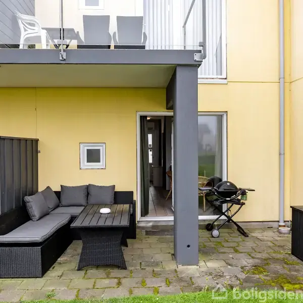 Terrasse med møbler i sort flet, et bord og en grill. Terrassen er delvist overdækket og ligger foran en gul bygning. Dør i baggrunden forbinder indendørs og udendørs.