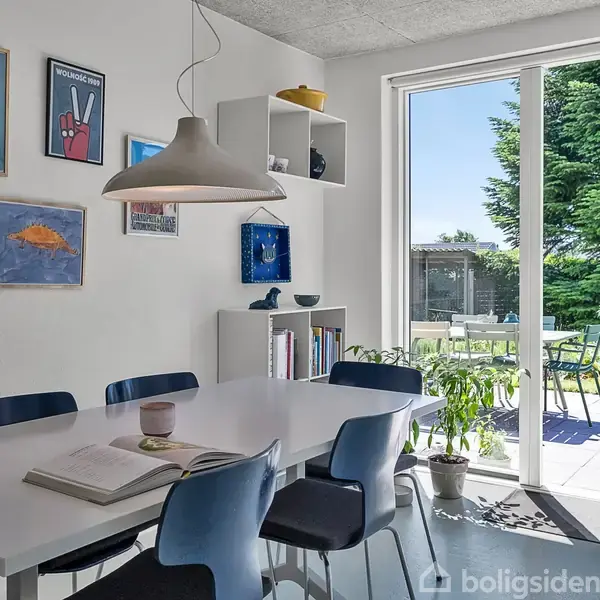 Et spisebord med blå stole står i et lyst rum. En åben dør fører ud til en solrig terrasse med grønne planter og havemøbler. Vægge har kunstværker.