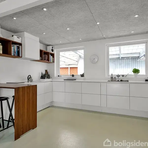 Et moderne køkken med hvide skabe og skuffer, hvor to sorte stole står ved en træbænk. Rummet er lyst op af dagslys fra to vinduer, og loftet har indbyggede spots.