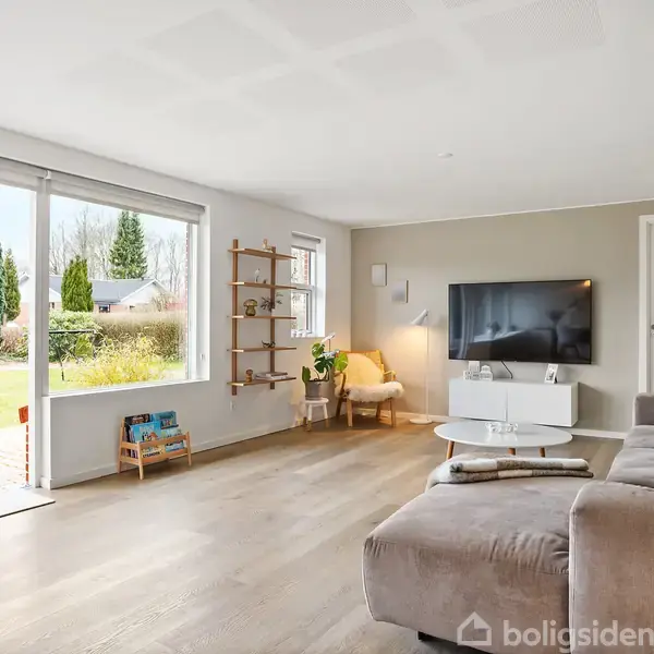 En lys stue med stor grå sofa står mod en væg med et fladskærms-TV. Store vinduer og glasdøre åbner ud til en have. En lænestol står i hjørnet.