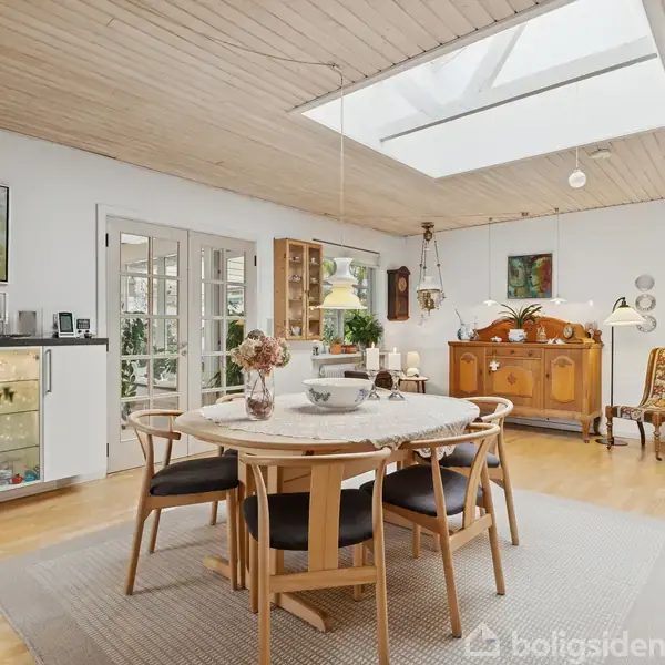 Rundt spisebord med seks stole står på et tæppe i et lyst rum med trægulv. Ovenlys i loftet. Væggen har kunst og skænke med dekorationer.