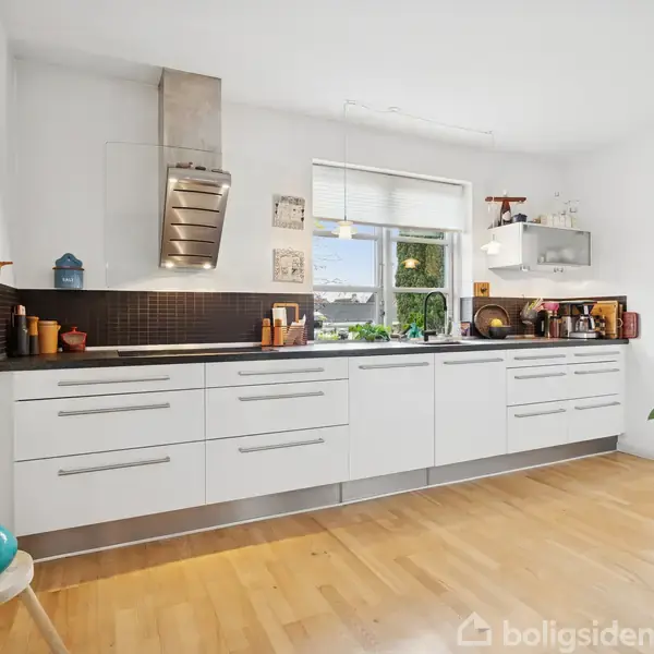 Hvidt køkken med lange skuffer og indbygget emhætte; planter og køkkenredskaber placeret på bordpladen. Trægulv og hvidmalede vægge med et stort vindue, der giver lys.