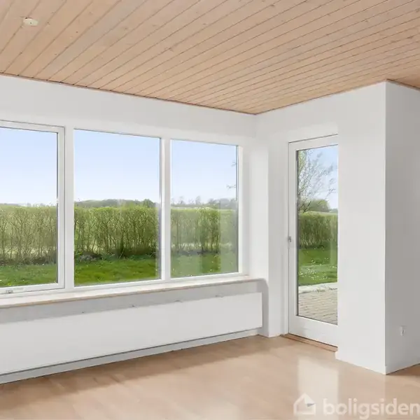 Stue med store vinduer og en glasdør, der giver udsigt til en grøn have med hæk. Lyse trægulve og træloft skaber en lys og indbydende atmosfære.