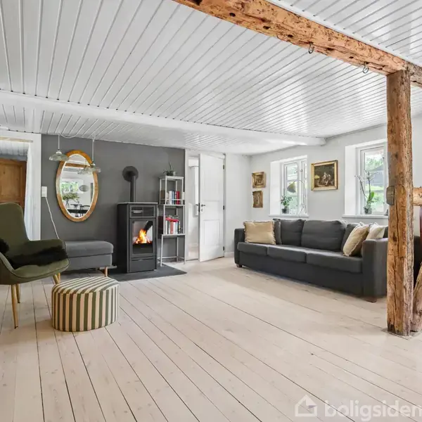 Stue med en grå sofa, en grøn lænestol, og en træbjælke som dekoration. Brændeovn ved grå væg skaber hyggelig stemning. Lyst trægulv og loftskonstruktion i træ.