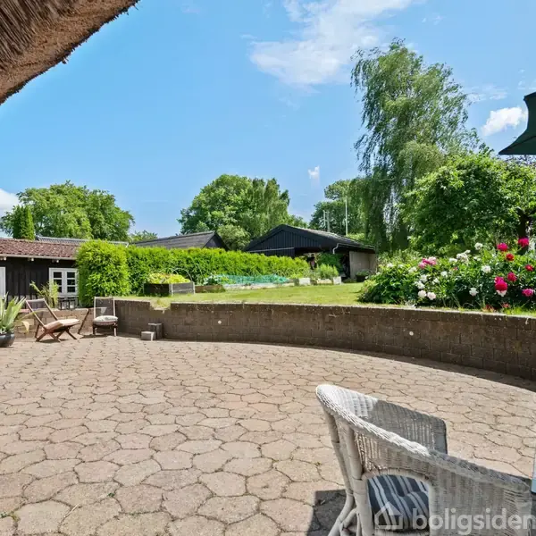 En rummelig terrasse med kurvemøbler og blomsterbede omkranset af en grøn have og træer under en klar blå himmel. I baggrunden ses en lav bygning med teglstenstag.