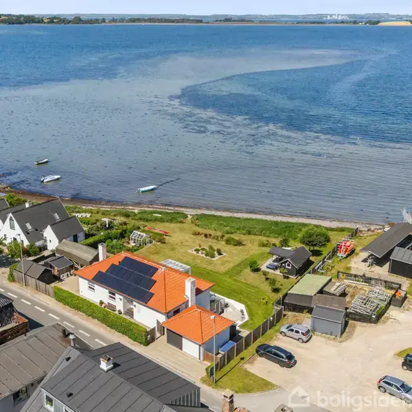 Huse langs kysten med en stor, blå bugt i baggrunden. Flere både ligger ved kysten, mens biler er parkeret foran boligerne. Grønt landskab omkranser området.
