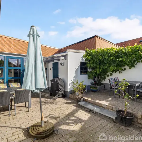 En brolagt gårdhave med havemøbler, en parasol og grill. Vægge med hvid mur og grønne planter pryder rummet. Solrigt med blå himmel i baggrunden.