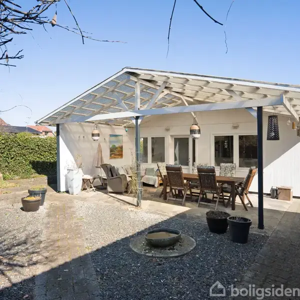 Overdækket terrasse med træmøbler og lamper under et hvidt tag. Beliggende i en stenbelagt gårdhave omgivet af grønne buske og en mur i baggrunden.