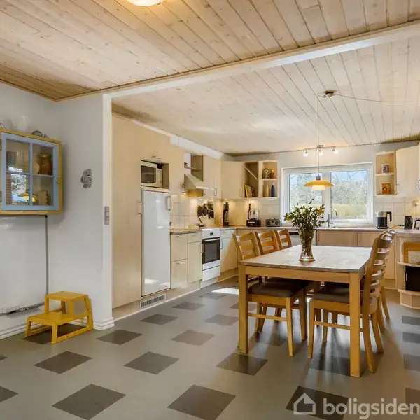 Køkken med spisebord og stole i midten, omgivet af hvidt køkkenskab og apparater. Store vinduer tillader naturligt lys. Trægulve og -lofter skaber en hyggelig atmosfære.