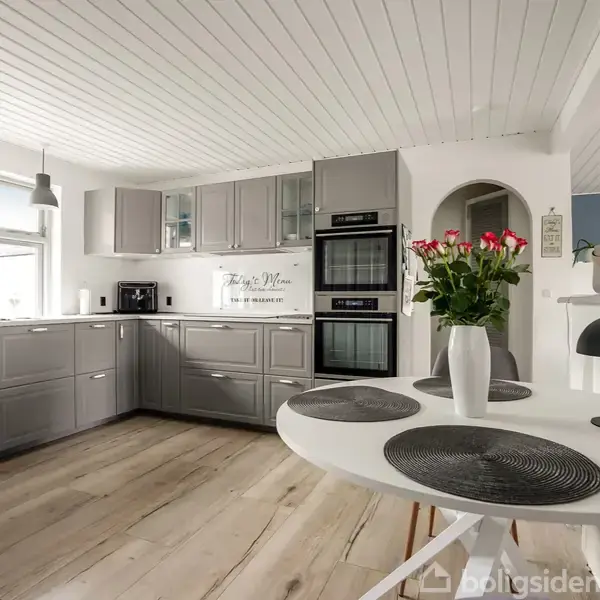 Et moderne køkken med grå skabslåger og to indbyggede ovne. På bordet står en vase med blomster. Rummet har trægulv, hvidt loft og store vinduer, der giver naturligt lys.