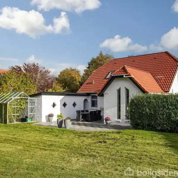 Et hvidt hus med rødt tag står i en frodig have. Til venstre er et drivhus placeret. Grønt græs og buske omgiver omgivelserne under en blå himmel.