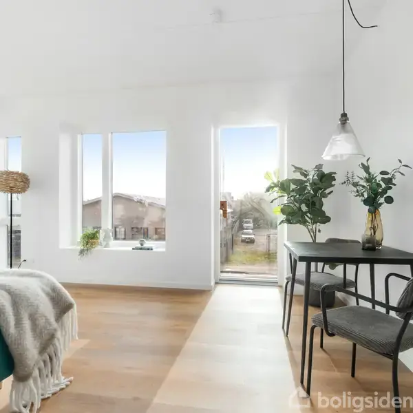 En lys stue indeholder et bord med planter, en stol og en seng dækket med et tæppe. Rummet har store vinduer og en dør ud mod en balkon.