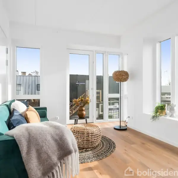 En grøn sofa med puder og tæppe står ved en lys stue. Rattanbord og plante på tæppe. Store vinduer og altandør giver udsigt til moderne boligkvarter.
