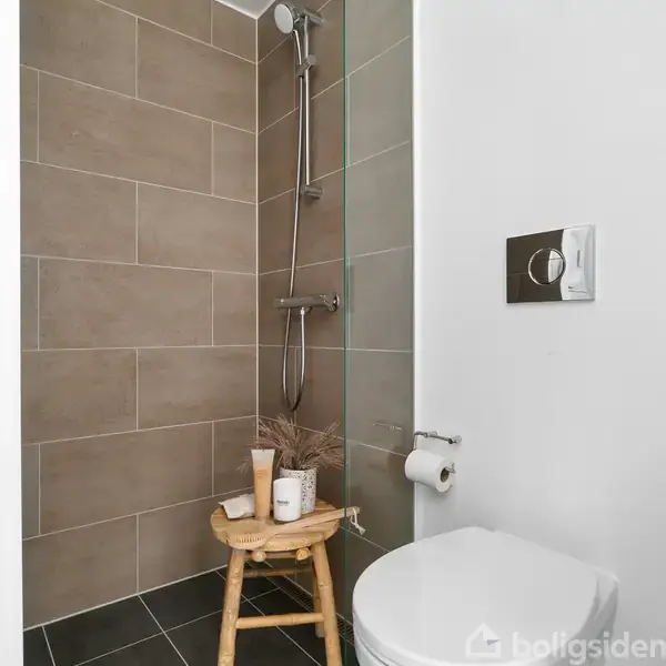 Bruser med håndbruser hænger på væggen i et badeværelse med brune fliser. En lille træskammel bærer toiletartikler og tørret græs ved siden af en toiletrulleholder.