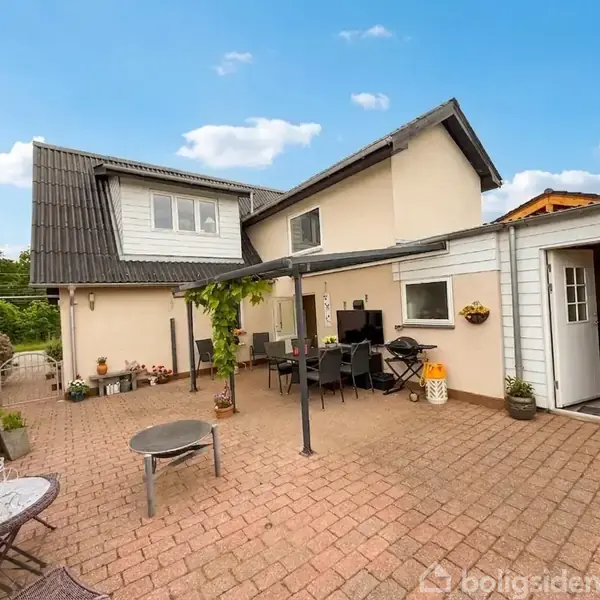 Et gårdområde med flisebelagt terrasse, hvor et bord- og stol-sæt står under en pergola. Vægdekorationer og blomsterpotter pynter husets udendørs vægge. Siden af huset ses i baggrunden.