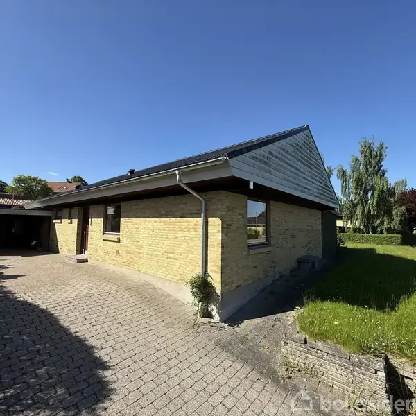 Et enfamilieshus af mursten står stille i et solbeskinnet boligområde. En brolagt indkørsel fører til en carport, og en græsplæne omgiver huset.