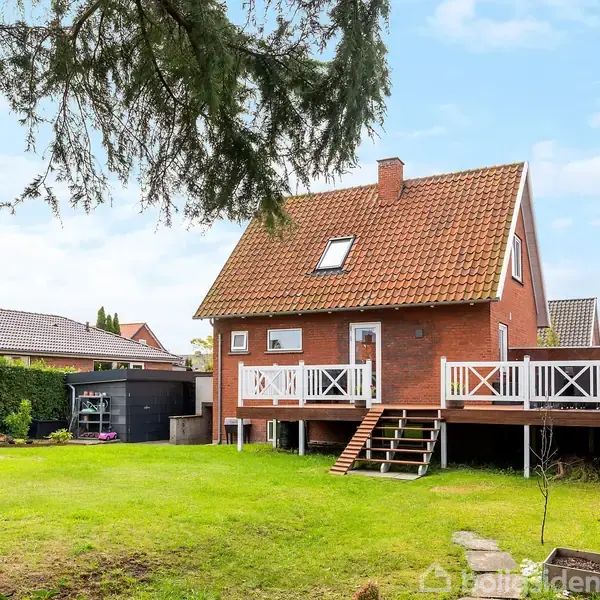 Et rødt murstenshus med saddeltag. Huset står på en stor græsplæne med en hævet veranda og trapper. Omkranset af grønne hække og træer under en blå himmel.