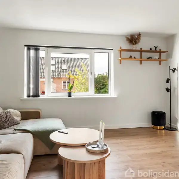 Stue med beige sofa, sofabord og lysestager opstillet foran et vindue. Væggene er hvide med en grøn accentvæg og forskellige dekorative elementer hænger oppe. En åben dør viser tilstødende rum.