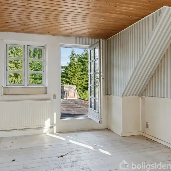 Et lyst rum har hvidmalede gulve og træloft, med en åben dør mod en terrasse. En radiator er placeret under et vindue, og træer ses udenfor.