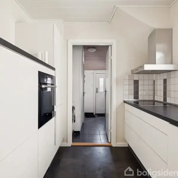 Et moderne køkken med hvide skabe og sorte bordplader, kombineret med en indbygget ovn. Der er fliser på væggen bag kogepladen og en stor vindue til højre.