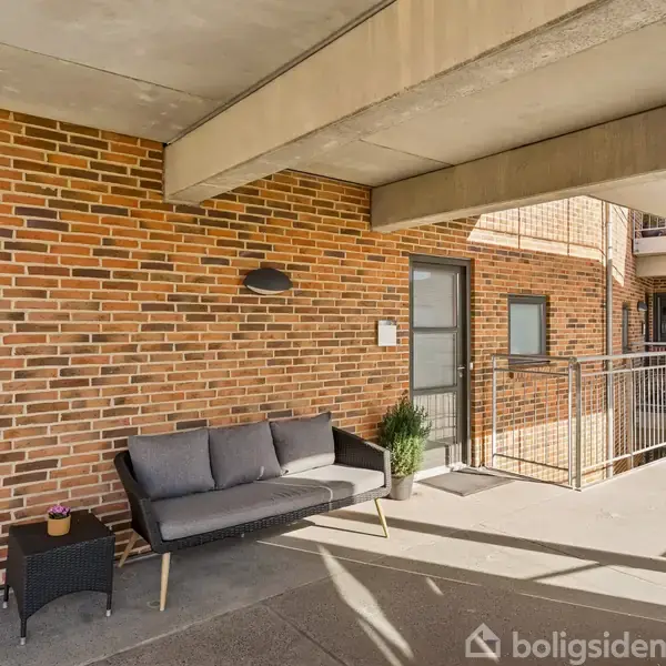 Sofa med grå hynder står på en balkon med røde murstensvægge. En sort bordlampe og grøn plante er ved siden af. Rækværk i metal giver udsigt til anden etage.