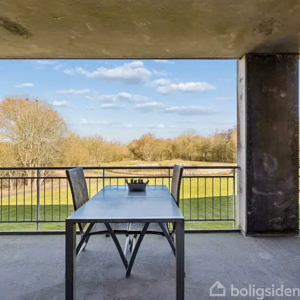 Bord og stole står ubevægeligt på en balkon med udsigt over en grøn, skovklædt park under en blå himmel.
