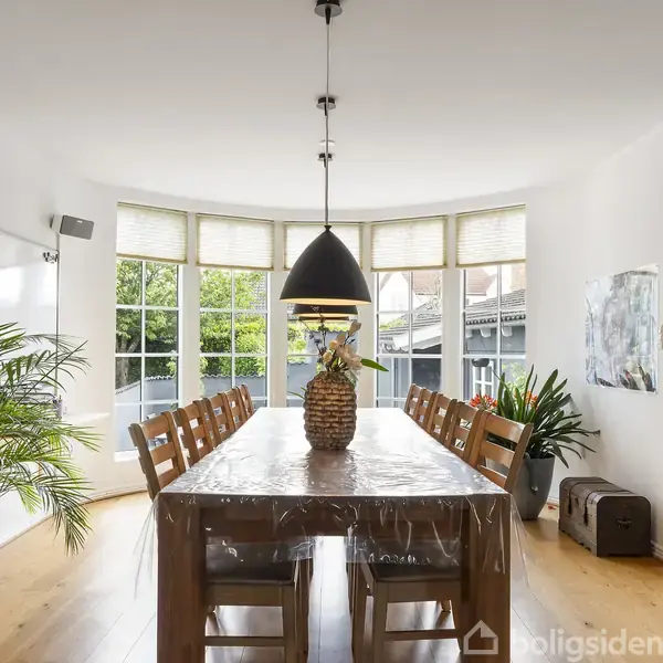 Spisebord med otte stole står under en sort pendellampe. Bordet er pyntet med en dekorativ krukke. Store vinduer bagved giver udsigt til et grønt område. Plante til venstre.