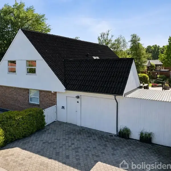 Huset står stille i et solrigt boligområde, omgivet af grønne træer og en flisebelagt indkørsel. Det har en sort tag og hvid facade med røde mursten nedenunder.