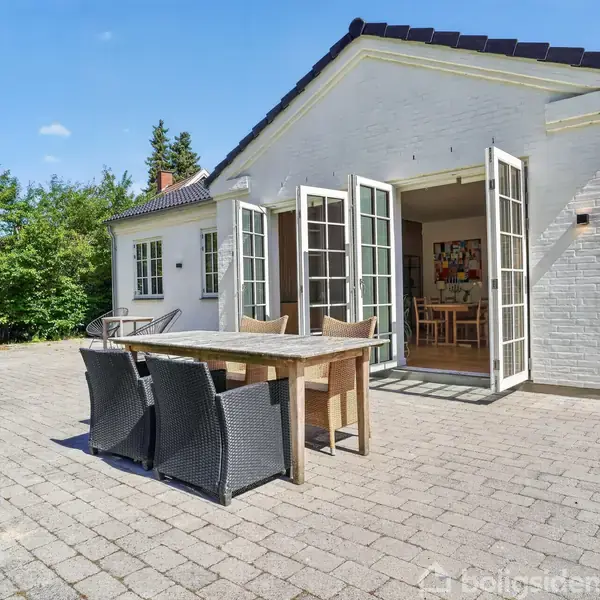 Et træbord står på en terrasse med fire stole omkring. Terassedørene er åbne til et hvidt hus. Omgivet af grønne buske og en græsplæne under en blå himmel.
