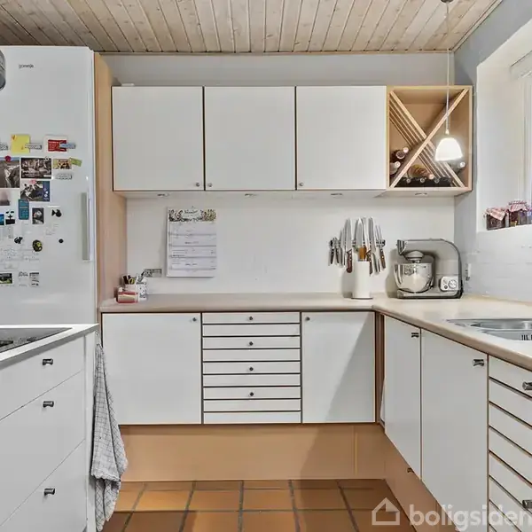 Hvidt køkken med moderne skabe er fyldt med forskellige køkkenredskaber, en ovn og et køleskab pyntet med magneter. Rummet har en lys træloft og vindue med udsigt.