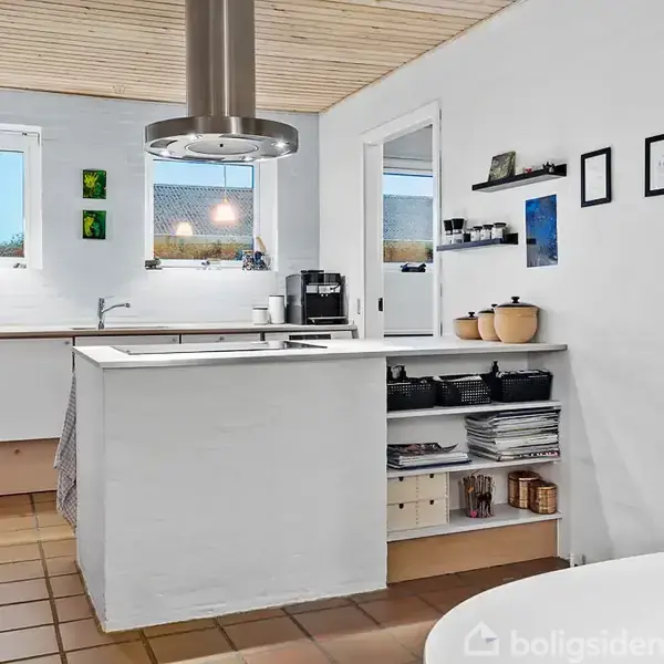 Et køkken med hvide vægge og træloft har en central køkkenø med hylder fyldt med køkkenudstyr og bøger. Der er også et gaskomfur og to vinduer med lamper.