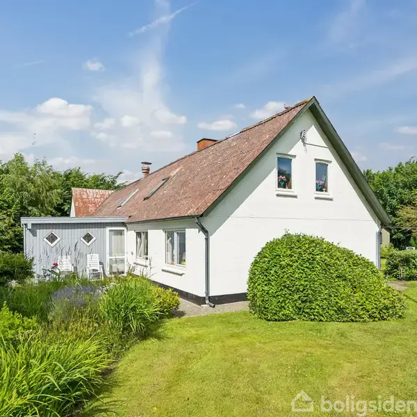 Et hvidt hus med rødt tegltag står stille omkranset af grønne buske og en velplejet græsplæne, under en blå himmel med skyer.