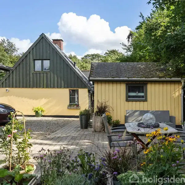 Hus malet i gule og grønne nuancer med stejl tagryg foran en parkeringsplads. Blomster fylder forgrunden, mens et bord med stole står i haven under blå himmel.