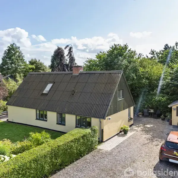 Et gult hus står i en have med grøn beplantning. En grusvej fører til en carport, hvor en rød bil er parkeret. Træer omringer området.