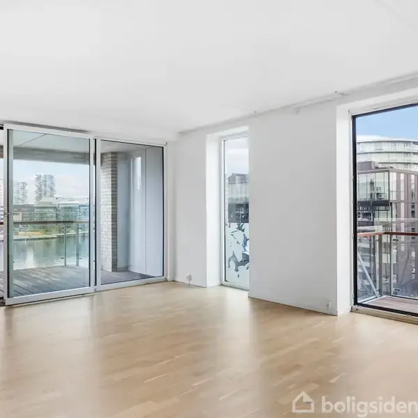 Stue med trægulv og store vinduer. Udsigt til moderne bygninger og balkon. Meget lysindfald gennem glasdøre.