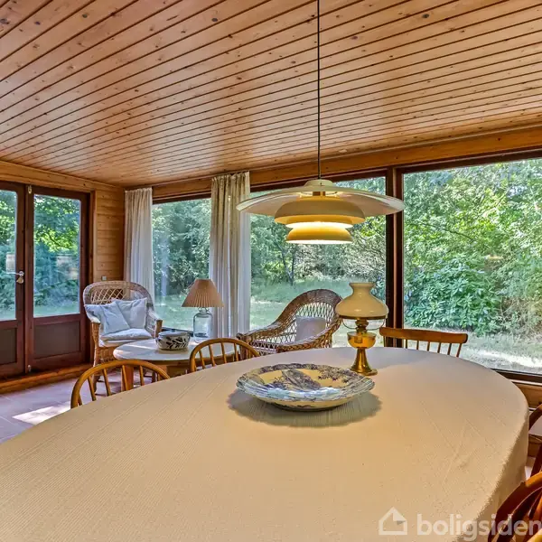 Langbord med dug står i centrum med stole rundt omkring. Rattan-møbler og lamper udfylder rummet. Omkring bordet er store vinduer, der giver udsigt til en frodig have.
